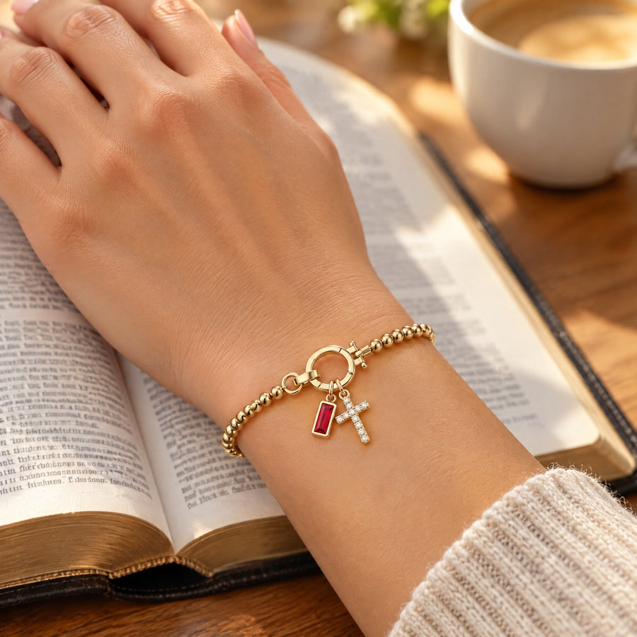 Gold cross charm birthstone bracelet on a woman's wrist, her hand resting on an open Bible