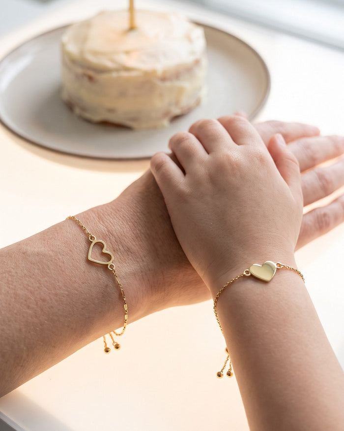 A daughter's hand resting on her mother's at a birthday table: open heart bracelet on the mother, filled heart bracelet on the daughter