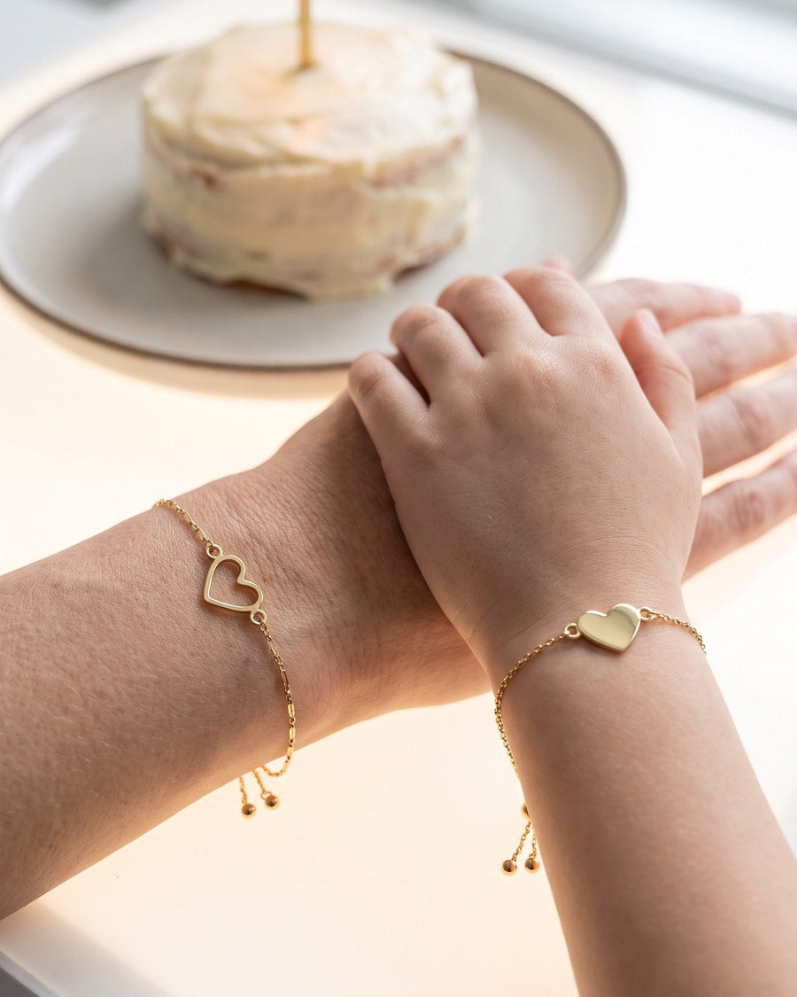 A daughter's hand resting on her mother's at a birthday table: open heart bracelet on the mother, filled heart bracelet on the daughter